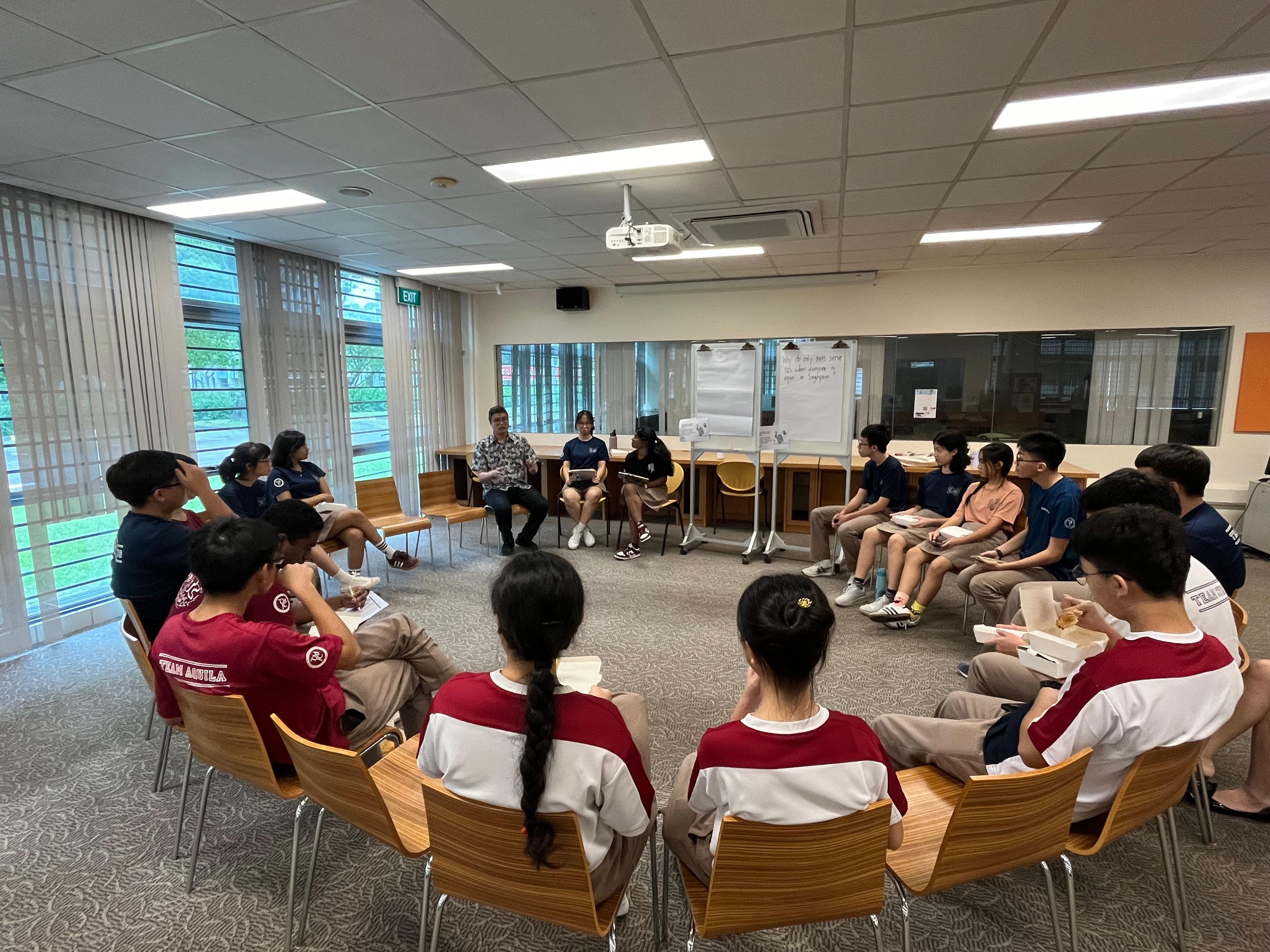 A group of about 15 students seated in a circle of chairs in a classroom, engaged in a discussion facilitated by two adults.